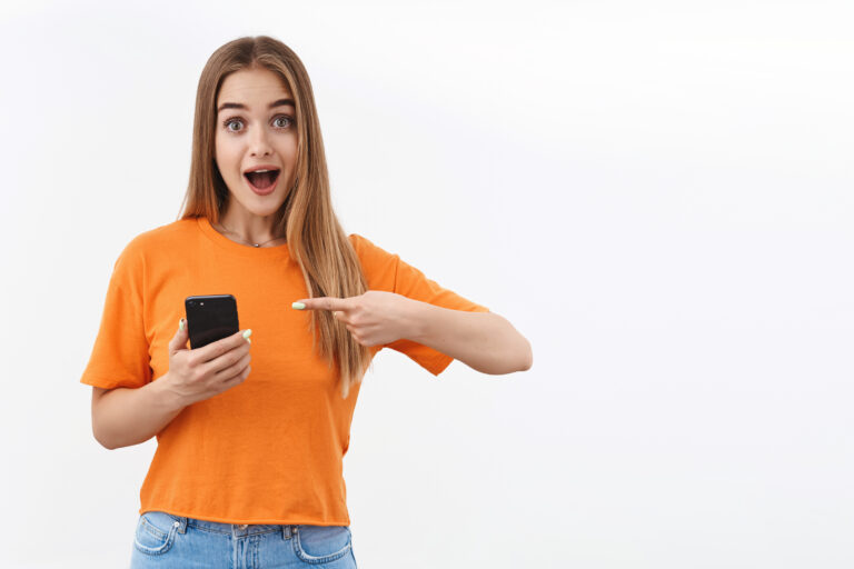 Technology, youth and communication concept. Portrait of excited, surprised blond girl pointing finger mobile phone, open mouth amazed, asking question about new app or online shopping site.